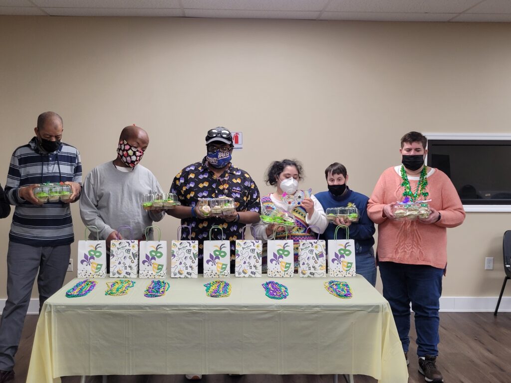 Six people standing behind a table, each holding a tray of cupcakes with colorful icing. On the table are mardi gras-themed bags and necklaces.