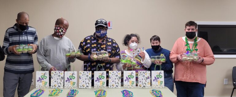 Six people standing behind a table, each holding a tray of cupcakes with colorful icing. On the table are mardi gras-themed bags and necklaces.