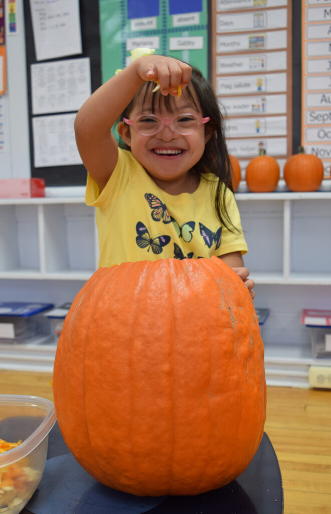 A girl in pink glasses and a yellow shirt smiling as she pulls pulp from a carved pumpkin.