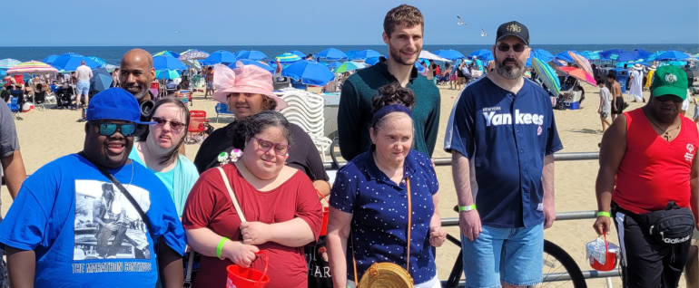 A group of nine people standing on the boardwalk with the beach and ocean behind them. The day is sunny and everyone is looking at the camera.