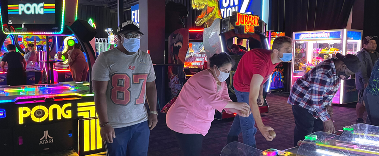 A group of four people playing skee-ball. More arcades and games are visible behind them.