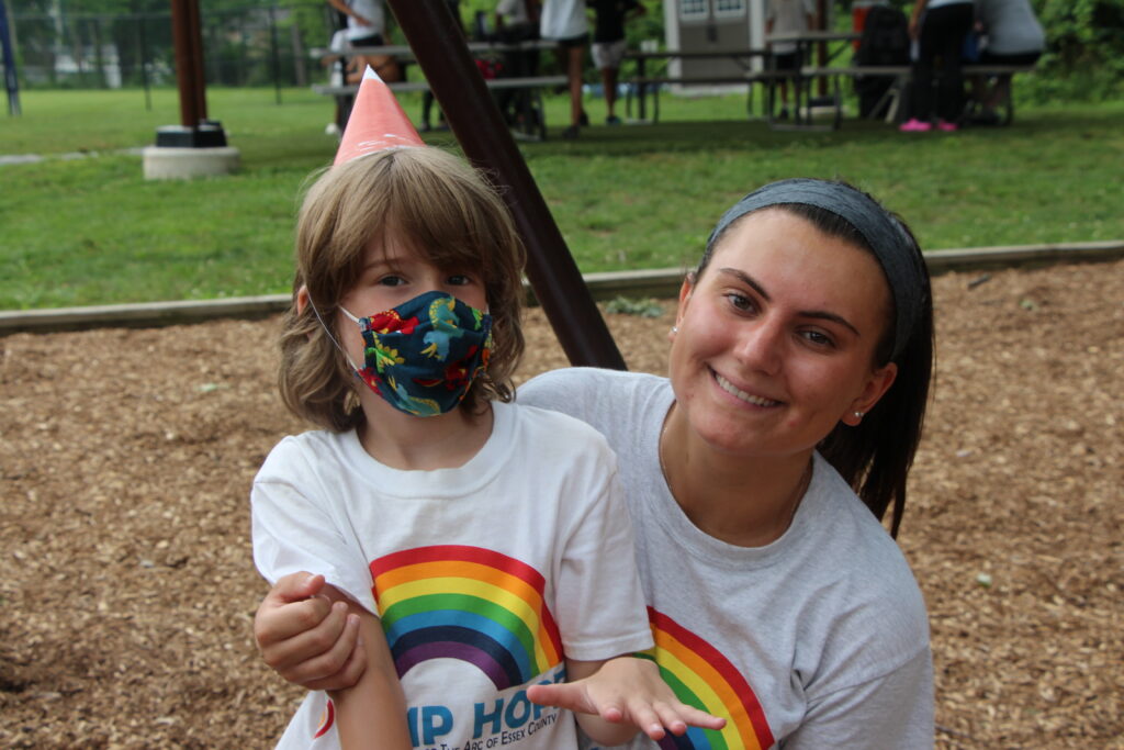 Camp Hope camper in a mask and a birthday party hat and counselor, smiling at camera
