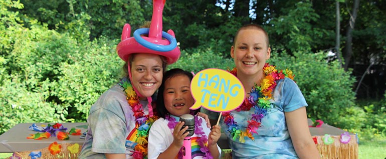 A girl sitting in between two women. All are wearing luau costumes (leis and bright colors). The woman on the left is wearing an inflatable flamingo hat. The girl in the center is holding a sign of a speaking bubble that says 