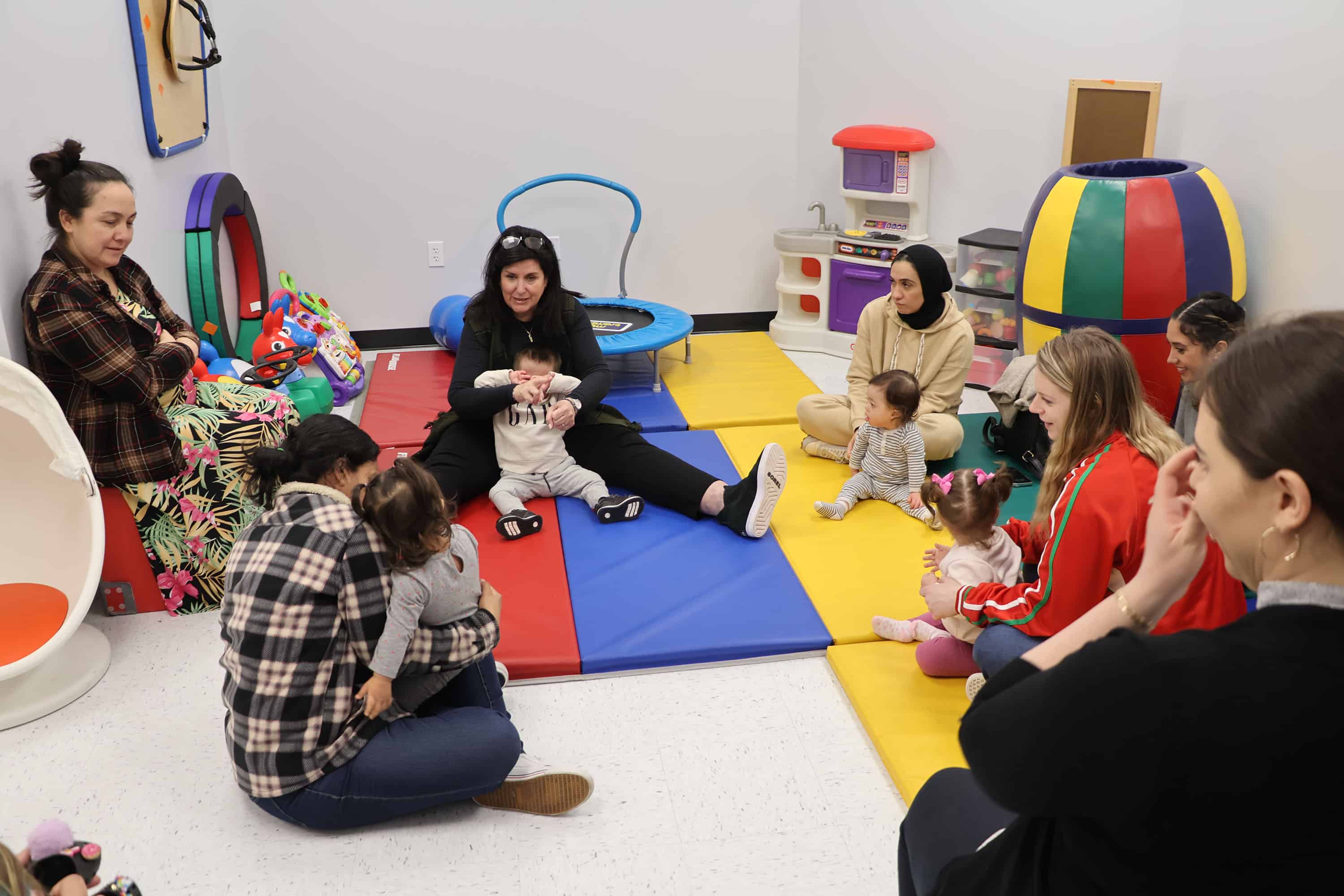 A group of adults with babies sit in a circle on a colorful mat, with therapy toys surrounding them.