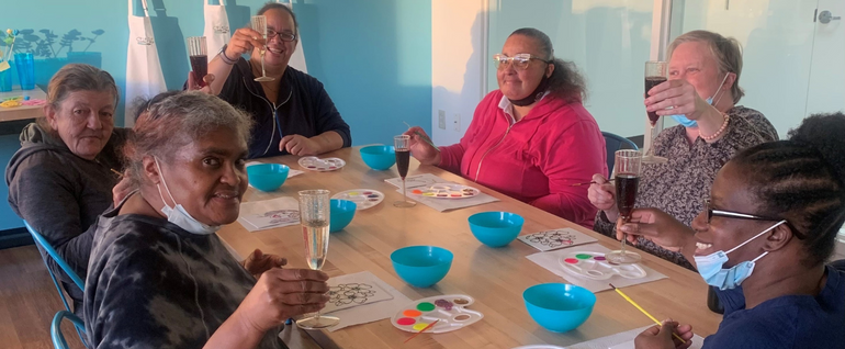 Six women sitting around a table, raising glasses, with a floral art project and paint in front of each of them.
