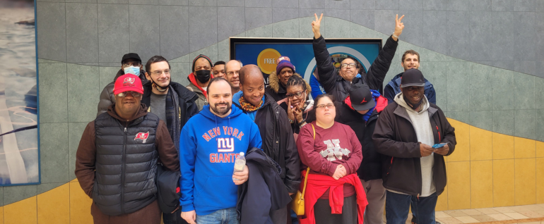A group of fifteen adults stand together in front of a wall that has different shades of blue and yellow tiling. One of the men in the group has both arms up in the air with peace signs.