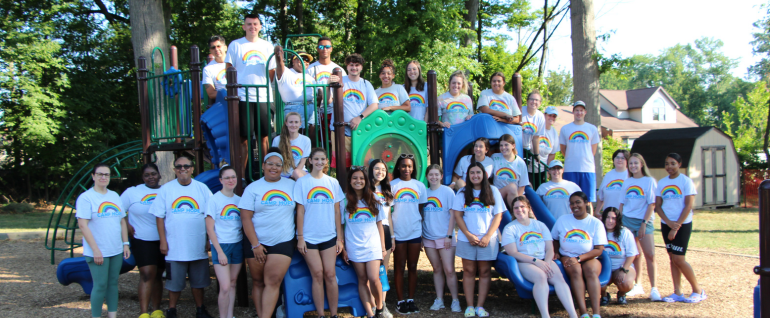 Camp Hope staff gathered on the Camp Hope playground for a group photo. All are wearing t-shirts with the rainbow Camp Hope logo on the front.