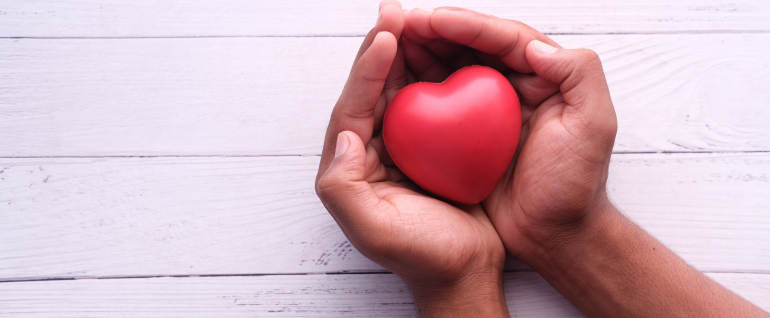 Two hands cupped together and holding a red heart object over a wooden white background.