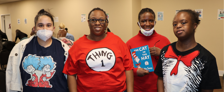 Four women wearing Dr. Seuss related shirts. The woman second from the right is holding the book "The Cat in the Hat."