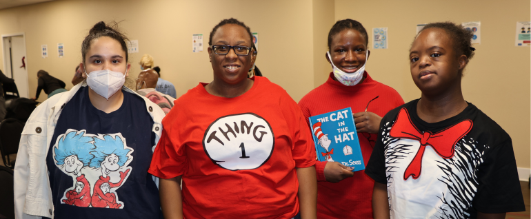Four women wearing Dr. Seuss related shirts. The woman second from the right is holding the book 