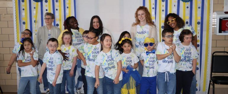 A group of students wearing shirts that say "Just Down Right Awesome," with several teachers and staff standing behind them. Blue and yellow streamer decorations are on the wall behind the group.
