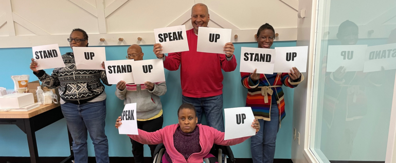 Five people holding signs that say "Stand Up" and "Speak Up." Four are standing along a wall, in front of them a woman sits in a wheelchair.