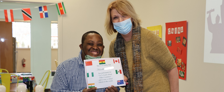 A young man and a woman stand next to each other. The man is holding a certificate and smiling. Behind them, decorations with different countries' flags are visible.