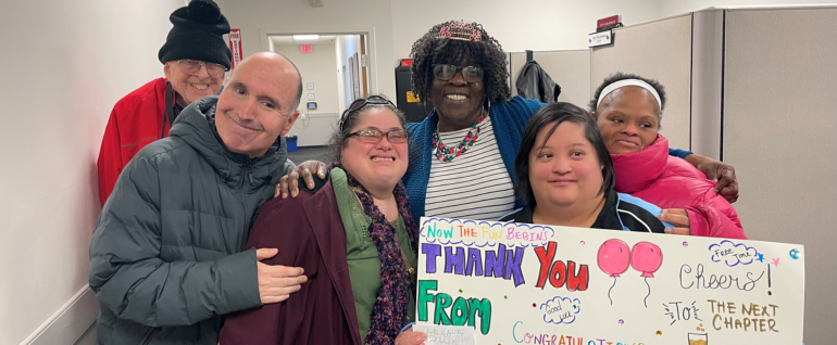 Six people across, embracing each other. Left to right: a man in a black hat and red jacket; a man in a dark gray coat; a woman in a green shirt with a black scarf and dark purple jacket, a woman in a blue and white striped shirt wearing a colorful necklace and a tiara, a woman in a black and blue shirt holding a poster board that says THANK YOU with balloons; and a woman in a pink jacket with a headband.