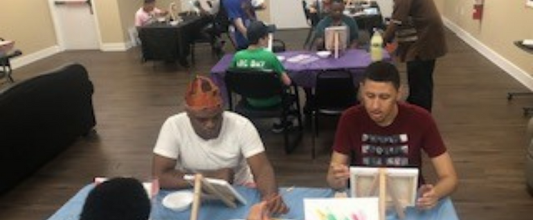 Two people sit at a table painting on canvases. More people are visible at tables in the background.