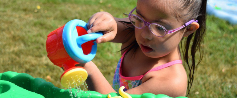 A girl in purple glasses and a bathing suit uses a toy watering can to pour water onto another toy.