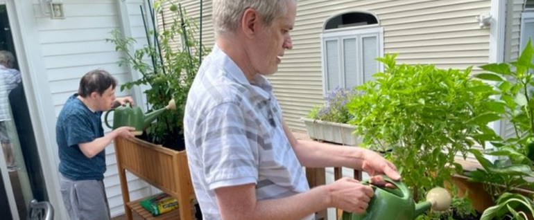Two men watering raised plant beds on a house deck.