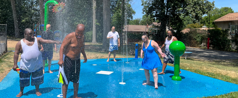 Six people in bathing suits standing amid the sprinklers on the splash pad at Camp Hope.