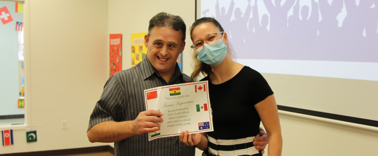 A man in a gray collared shirt holds up a certificate with different countries' flags on it while smiling next to a woman in a black shirt and blue mask.