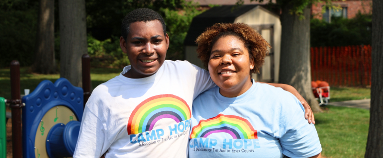 Two people stand in front of a playground. Both are wearing Camp Hope t-shirts and smiling at the camera.