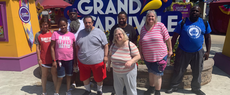Eight people standing in front of a sign that says "Grand Carnivale" on a sunny day.