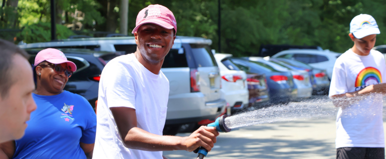 A young man in a red baseball cap stands in a sunny parking lot squirting a hose and smiling at the camera.