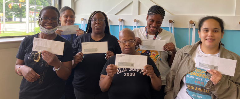 Six women stand together in front of a window and a blue and white wall, each holding an envelope up with a check inside.