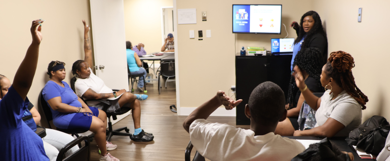 In a room with cream colored walls, a woman stands in the front at a monitor, showing a slideshow about financial literacy. You can see several people gathered around the edges of the room, several of whom are raising their hands.