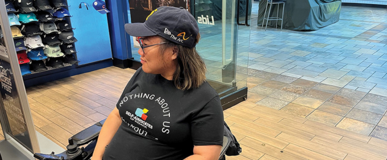 A woman with short brown hair sitting in a Lids hat store, wearing a black hat with The Arc logo on the side.