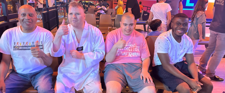 Four men sitting together in a row of seats at a bowling alley. The lighting has a reddish glow. All four are smiling at the camera, and three are giving a thumbs up.