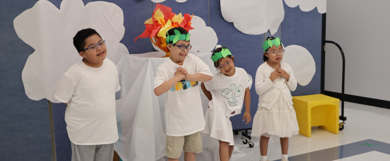 Four children wearing white stand together with green paper leaf crowns and a "torch" made of red, orange, and yellow paper behind them.