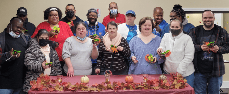 A group of 15 adults holding handmade crafts and standing behind a table decorated for Thanksgiving.