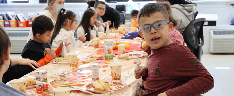A boy sitting at a table at school, where the children are all eating Thanksgiving food. The boy looking at the camera is wearing a red shirt and blue glasses.