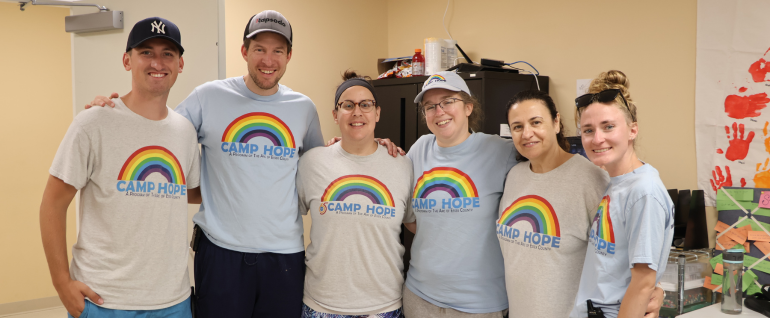 Six adults gathered together and smiling, all wearing Camp Hope t-shirts.