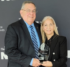 A man and a woman stand together, holding a glass award.