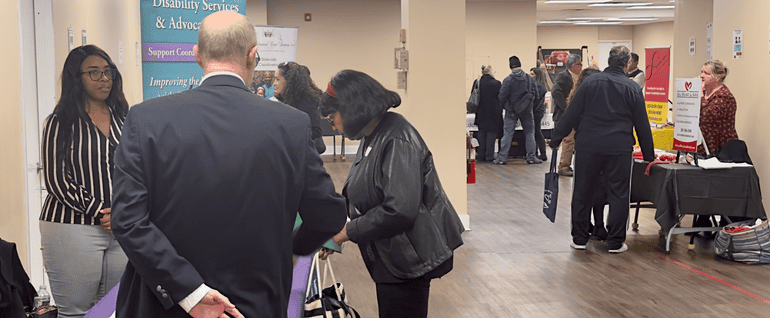 People mingling and stopping at tables to talk to representatives at different organizations. The tables have displays and printed materials spread out.