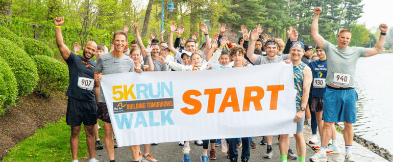 A group of runners stands behind a Building Tomorrows starting line banner, with trees on the left and a reservoir visible on the right.
