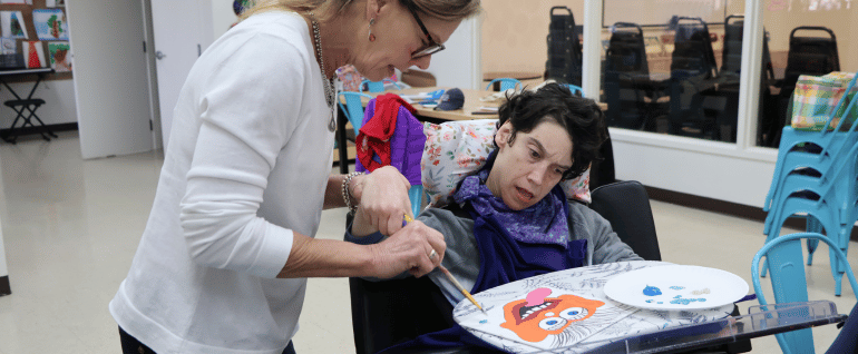 A woman standing, helping a woman in a wheelchair paint an orange monster face on canvas.