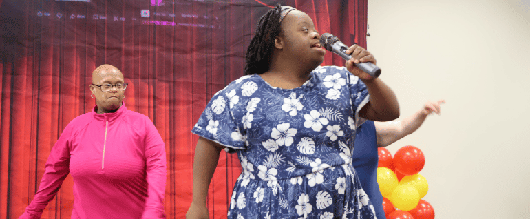 A woman in a blue dress with white flowers, singing into a microphone, with a woman in pink standing behind her and a red curtain backdrop.