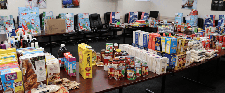 A collection of donated food items on a table, with full holiday gift bags visible in the background.