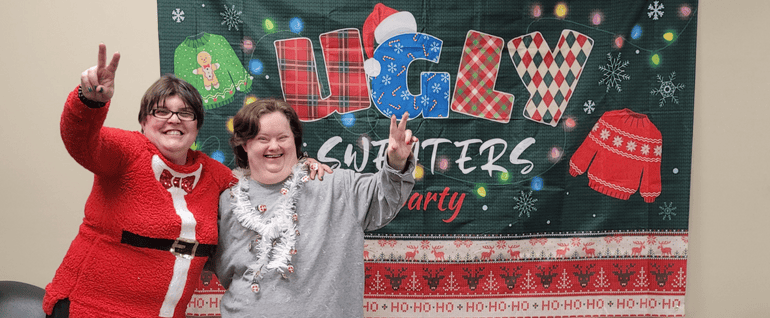 Two women leaning together, each holding up a peace sign. They are wearing holiday attire, and the decoration on the wall behind them says 