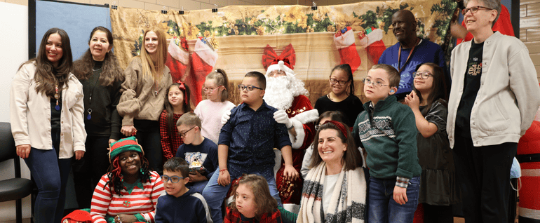 A group of children and staff gathered around Santa Claus with a holiday theme background behind them.