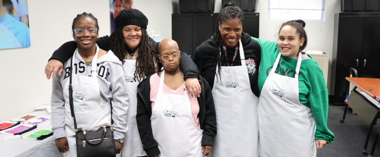 A group of five women stand together wearing white Studio Arc aprons.