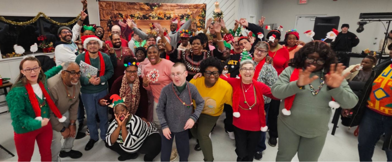 A large group of adults, many wearing holiday clothing and standing together in front of a holiday scene backdrop.