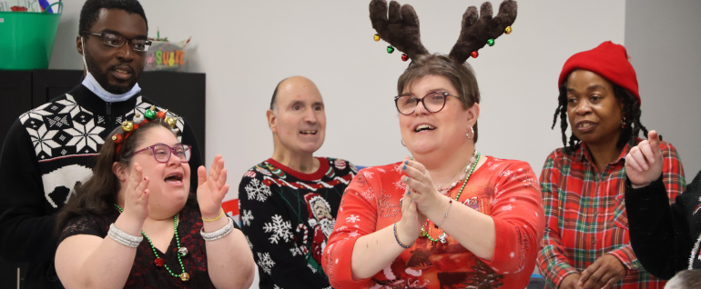 A group of five adults wearing holiday clothing and singing and clapping together.