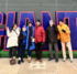 A group of six people stand in front of a large cutout of the word GIANTS with green turf underneath, in front of MetLife Stadium.