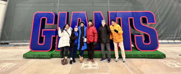 A group of six people stand in front of a large cutout of the word GIANTS with green turf underneath, in front of MetLife Stadium.