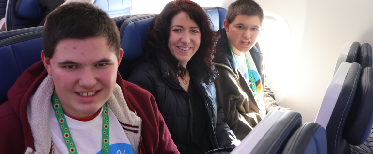 A woman sits in between two teenage boys in an airplane row on a United aircraft. The boys are wearing sunflower lanyards and their Wings for All t-shirts.