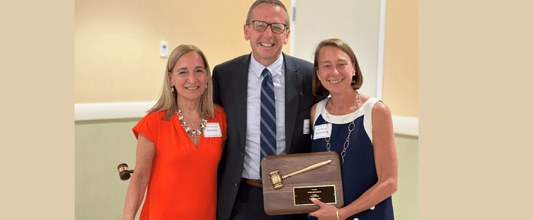 Three people stand together: A woman in a red dress, a man in a suit, and a woman in a navy and white dress. The woman on the right is holding a plaque with a gavel on it.