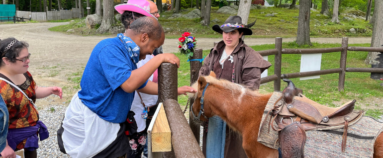 A man stands by a fence and pets a horse. Two other people stand on the side of the fence with him. On the other side of the fence, near the horse, is a woman in western gear.